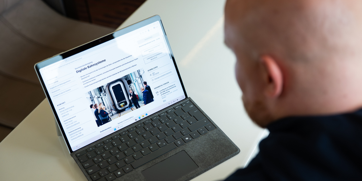 Bild eines aufgeklappten Laptops auf dem die Webseite des Studiengangs Digitale Bahnsysteme der HSBI zu sehen ist Im rechten Bildrand ist der Hinterkopf von Johannes Reinholz zu sehen
