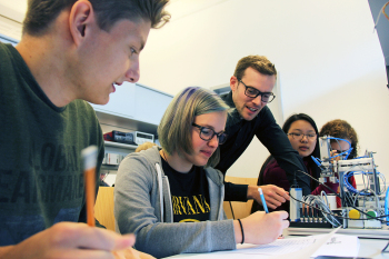 Die Schüler sitzen am Tisch mit dem Greifarm und Wilmsmeyer beicht sich zu ihnne hinunter.