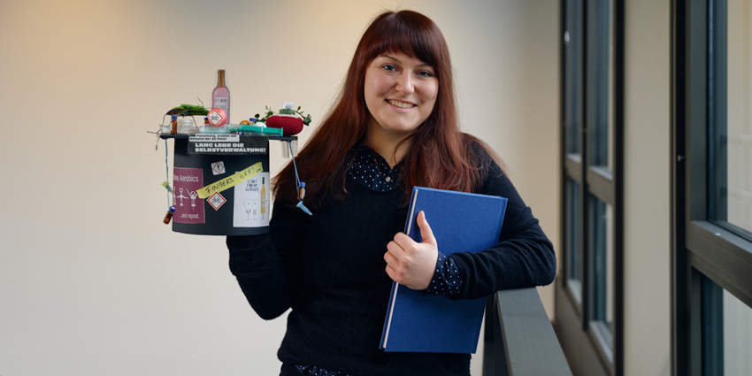 Linda Muskat im Porträt Sie hält ihre Doktorarbeit sowie einen bunten selbstgebastelten Doktorhut in die Höhe