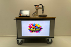 Foto: Der Pancake-Roboter PanCasso von vorne mit Logo auf dem Bildschirm und dem Roboterarm über dem Pfannkuchen-Eisen