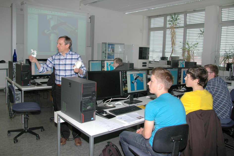 Der Dozent hält ein frisch gegossenes Plastik-Objekt in die Höhe. Drei Schüler sitzen vor Computern und betrachten das Objekt interessiert.