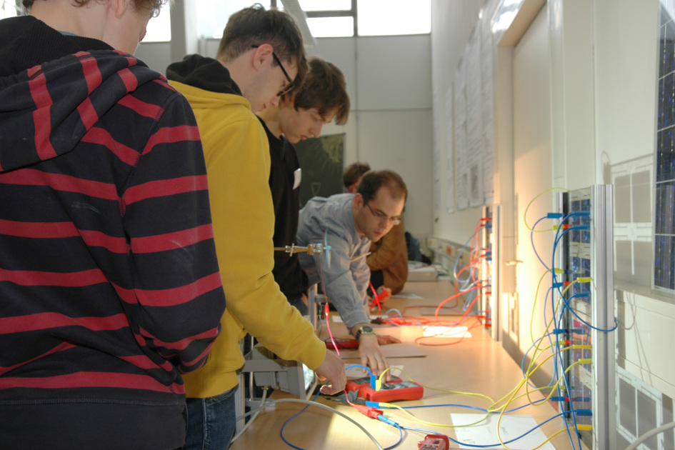 Drei Schüler verbinden mit Kabeln eine Solarzelle mit einem Stromumwandler. Ein wissenschaftlicher Mitarbeiter zeigt ihnen, wie das geht.