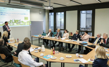 Ein Mann vor einer Leinwand in einem Seminarraum begrüßt anwesende Personen die im Halbkreis an Tischen sitzten und ihm zuhören.