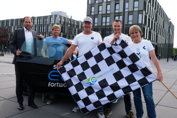 Fünf Personen stehen vor einem Auto und halten eine karierte Fahne. .