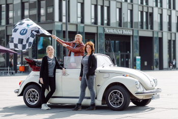 Eine Person steht in einem Käfer-Cabrio und schwinkt eine Fahne. Zwei Personen stehen daneben.