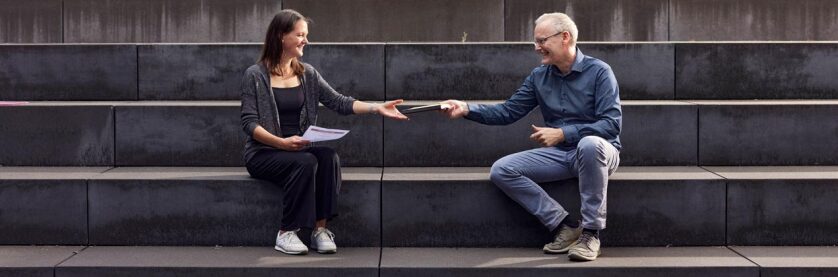Ein Mann und eine Frau sitzen auf einer Steintreppe und reichen sich ein Buch