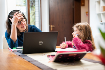 Eine Person sitzt an einem Tisch vor einem Laptop, neben ihr sitzt ein Kind und malt.