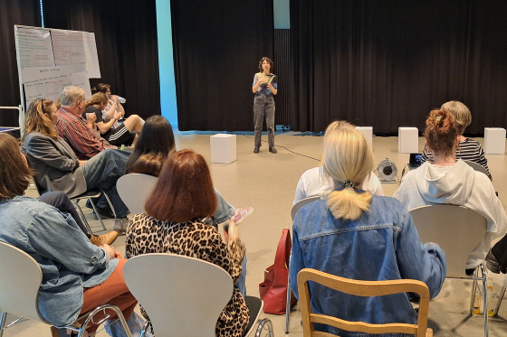 Foto: Mehrere Menschen sitzen in einem Stuhlkreis im Theater der HSBi und hören einer Professorin zu