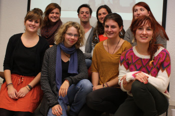 8 Studierende lächseln hockend in bunter Kleidung in die Kamera.