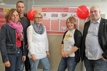 Die beiden Studireenden stehen rechts und links neben ihrem Poster. Hinter ihnen stehen die Dozenten des Studiengangs, zwei links, einer rechts.