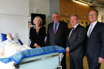 Vier Personen stehen neben einem Pflegebett, in dem eine menschengro0e Puppe liegt.