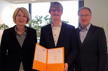 Zwei Männer und eine Frau stehen nebeneinander und der Mann in der Mitte hält eine Urkunde in die Kamera