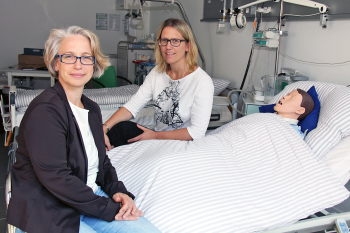 Prof. Dr. Michaela Brause und Sandra Tewes sitzen auf einem Krankenhausbett.