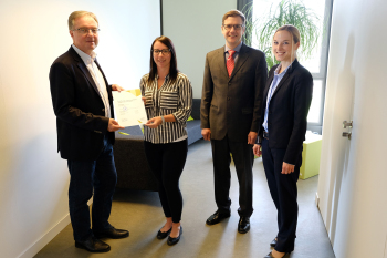 Vier Personen stehen im Flur, zwei Personen halten gemeinsam ein Blatt Papier in den Händen.