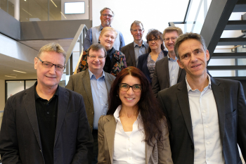 Drei Frauen und 6 Männer stehen nebeneinander und hintereinander auf einer Treppe und schauen in die Kamera.