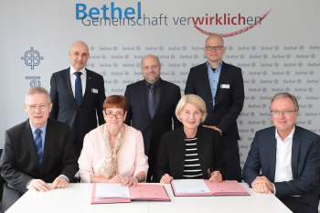 Zwei Männer und zwei Frauen sitzen an einem Tisch, dahinter stehen drei Männer