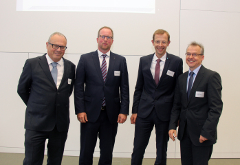 Vier Männer in Anzügen stehen vor einer Wand und schauen in die Kamera