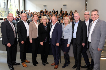 Neun Personen stehen nebeneinander und blicken in die Kamera im Hintergrund ist ein Hörsaal zu sehen.