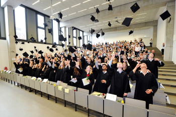 Eine Gruppe Menschen steht in einem Hörsaal und wirft Doktorhüte in die Luft