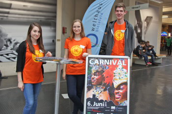 Die drei Studierenden stehen neben einem Aufsteller vom Carbival der Kulturen und einem Wimpel vom Welthaus.