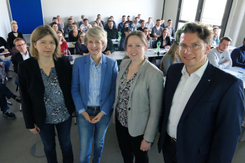 Die vier Personen stehen im Seminarraum vor den Studierenden.