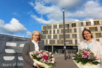 Zwei Frauen mit Blumensträußen stehen vor dem Hauptgebäude der FH Bielefeld und lächeln in die Kamera.