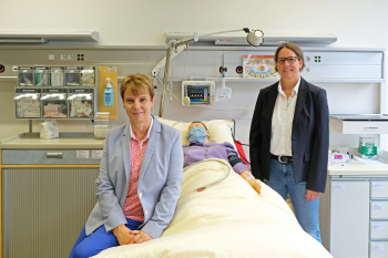 Eine Frau sitzt seitlich auf einem Krankenhausbett, in dem eine menschliche Puppe liegt. Rechts vom Bett steht eine weitere Frau. Im Hintergrund erkennt man medizinisches Zubehör.