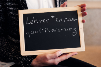 In der Hand wird eine Tafel mit der Aufschrift „Lehrer*innenqualifizierung“ gehalten