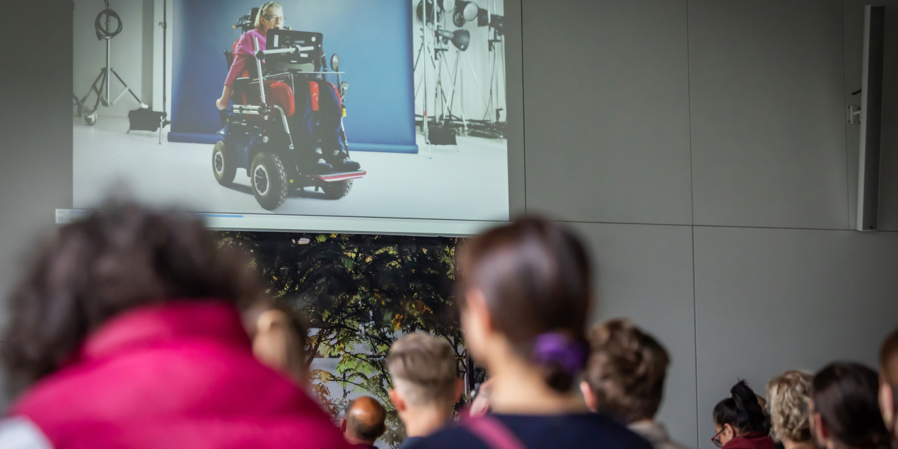 Auf der Leinwand im Konferenzbereich ist ein Film zu sehen. Die Szene zeigt eine Frau, die im Rollstuhl sitzt.