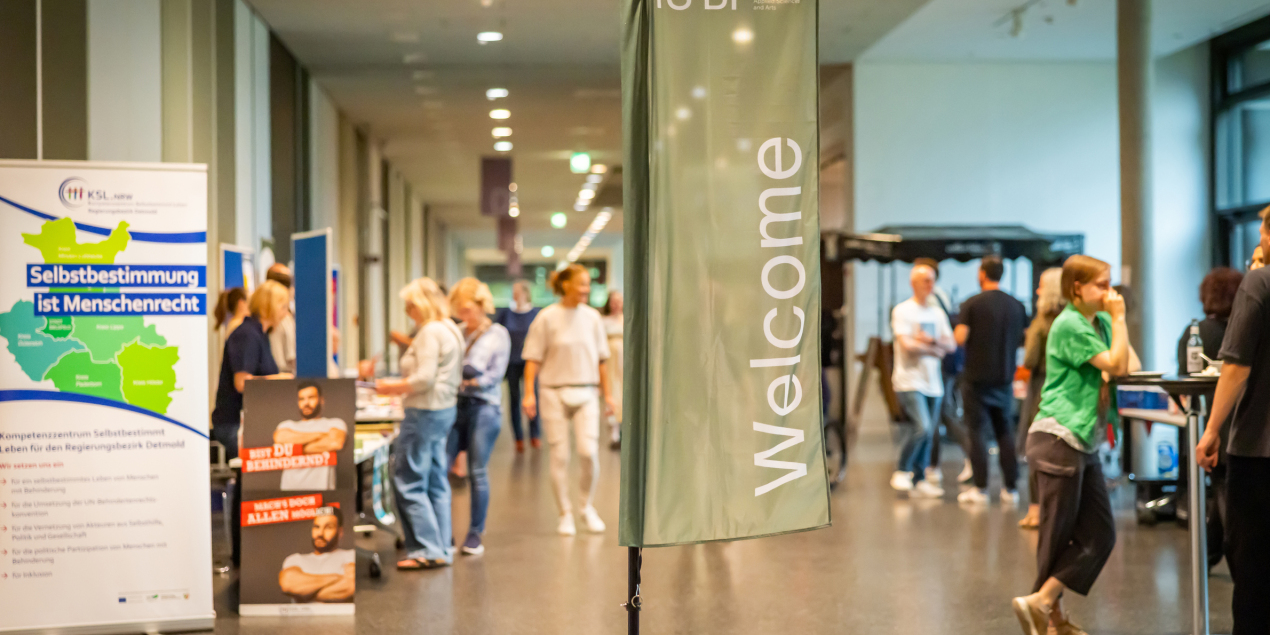 In der Magistrale der HSBI steht eine große Flagge mit der Aufschrift Welcome. Im Hintergrund sind Menschen und Infostände zu sehen