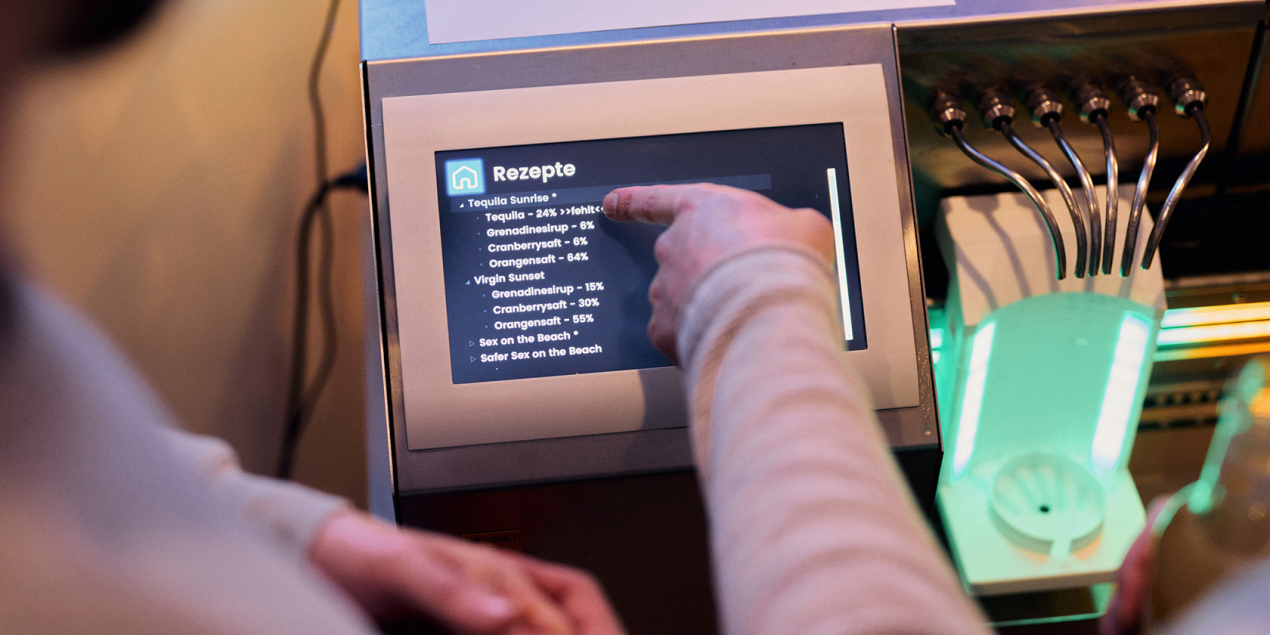 Eine Person zeigt auf das Bedienfeld des Cocktailautomaten mit der Rezeptauswahl