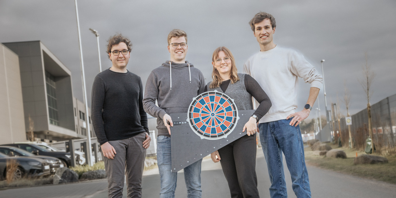 Gruppenbild des Entwickler innen Teams von links nach rechts sieht man Pierre Louis Detemble Felix Lütkebohle Jule Brede und Linus Bröker die auf dem Parkplatz des Projektpartners Beckhoff stehen und ihre Dartscheibe präsentieren
