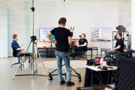 Im Hintergrund sitzen drei Personen in einem Kreis sich gegenüber, im Vordergrund sieht man einen Mann vor einer Kamera.