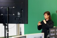 Im Vordergrund sieht man die Regie für die Aufnahme des Live-Streams. Vor einem Greenscreen steht eine Gebärdendolmetscherin.