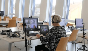 eine Person sitzt an einem Arbeitsplatz vor zwei Bildschirmen, auf denen man Teilnehmerinnen und Teilnehmer einer Videokonferenz sieht