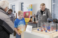 Ein Junge und ein Mann stehen vor einem Tisch auf dem ein Roboterarm und Säulen mit roten und blauen Kugeln zu sehen sind