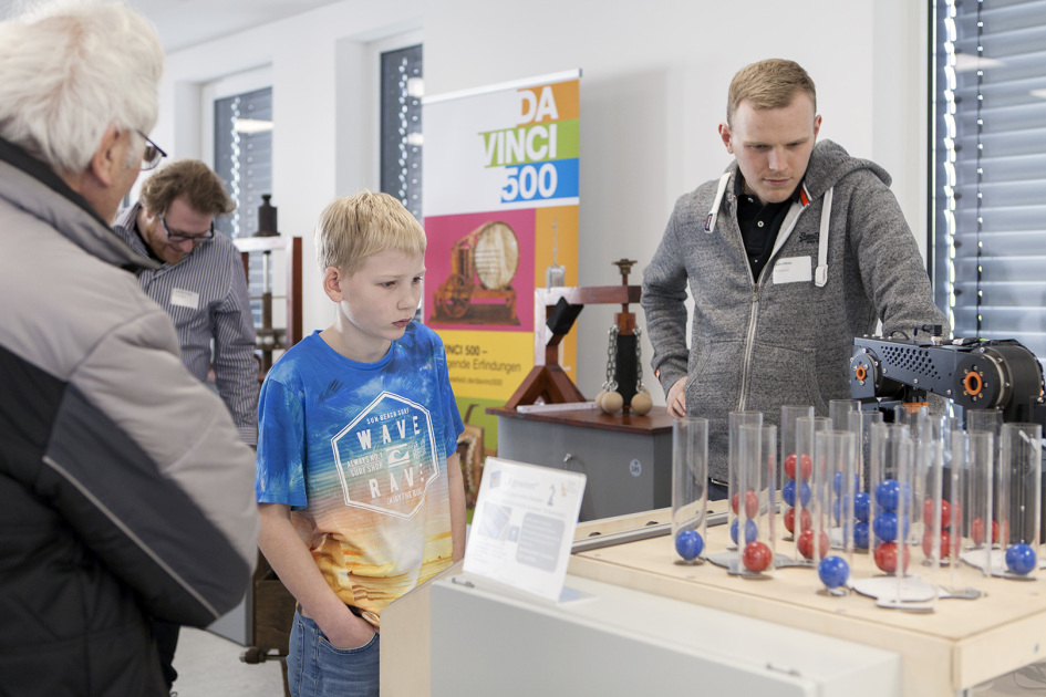 Ein Junge und ein Mann stehen vor einem Tisch auf dem ein Roboterarm und Säulen mit roten und blauen Kugeln zu sehen sind