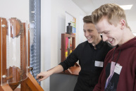 Zwei Männer stehen vor einer Gerüst aus Holz, einer der beiden dreht an einer Kurbel