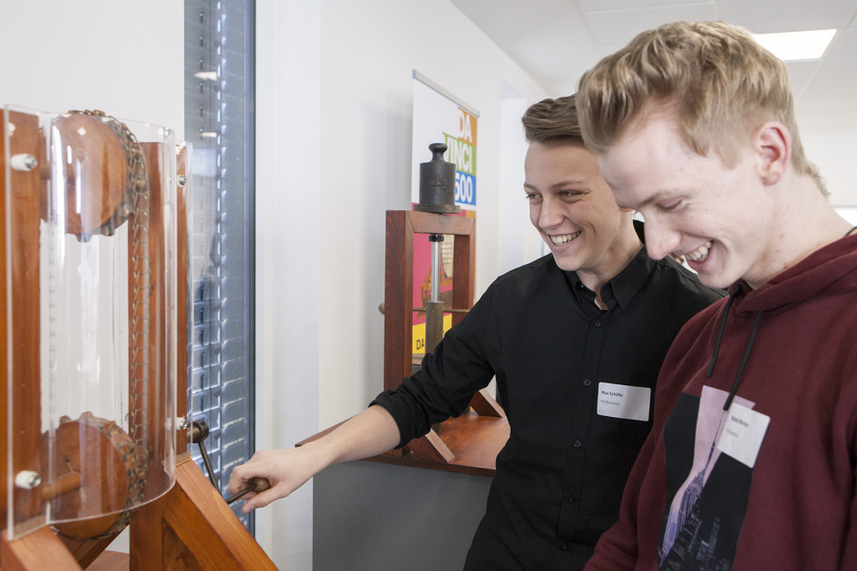 Zwei Männer stehen vor einer Gerüst aus Holz, einer der beiden dreht an einer Kurbel