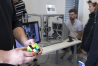 Im Vordergrund halten zwei Hände einen Zauberwürfel und im Hintergrund sitzt ein Mann vor einem Bildschirm