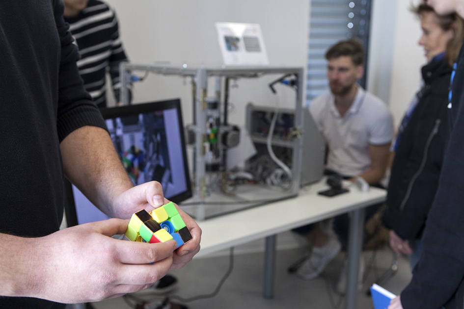 Im Vordergrund halten zwei Hände einen Zauberwürfel und im Hintergrund sitzt ein Mann vor einem Bildschirm