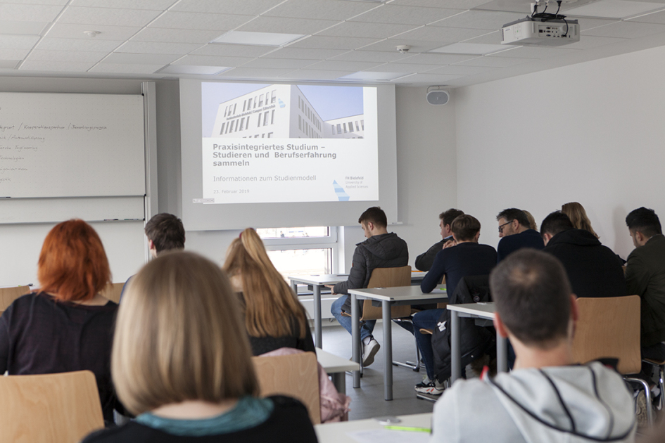 Eine Gruppe Menschen sitzt in einem Seminarraum vor einer Leinwand mit der Überschrift Praxisintegriertes Studium