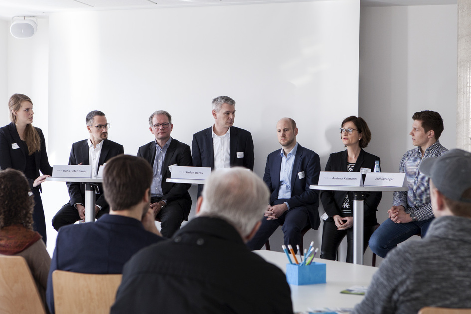 Zwei Frauen und fünf Männer sitzen vor einer Gruppe Menschen