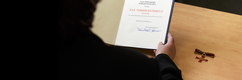 Barbara Schwarze blickt auf die Urkunde, auf dem Tisch liegt das Bundesverdienstkreuz