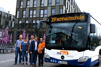 Vier Personen, zwei weiblich und zwei männlich, stehen links neben einem Bus. Im Hintergrund ist das Hauptgebäude der FH Bielefeld zu sehen.