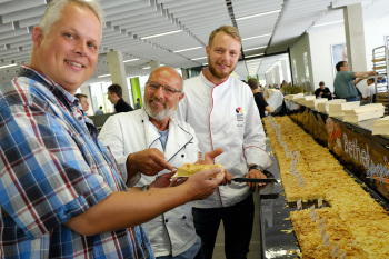 Drei Männer stehen vor einem Tablett mit Kuchen. Die Person in der Mitte übergibt der Person links ein Stück Kuchen auf einer Serviette. Die Person rechts steht daneben.