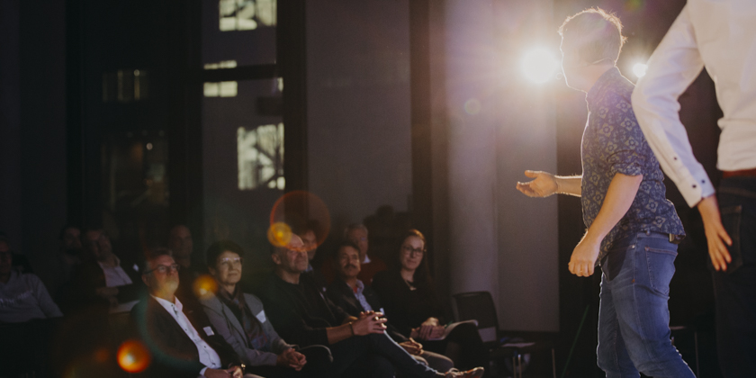 Auf der abgedunkelten Bühne im Konferenzsaal der HSBI stehen Marvin Meinold und Ben Hartwig Meinold trägt ein blaues geblümtes Hemd und ein Headset Hartwig steht in einem weißen Hemd und blauer Jeans rechts neben ihm auch er trägt ein Headset
