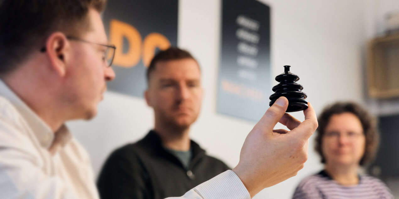 Drei Personen sitzen gemeinsam an einem Tisch und tauschen sich aus. Ein Mann hält einen schwarzen kleinen Faltenbalg in die Kamera.