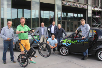 Vertreter der FH Bielefeld, Stadtwerke Bielefeld und e-CROSS GERMANY posieren mit Elektrofahrzeugen vor dem FH-Haupteingang.