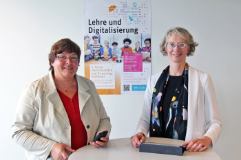 Prof. Dr. Tobina Brinker und Dr. Karin Ilg stehen mit digitalen Medien vor dem Plakat zur E-Learning-Konferenz\